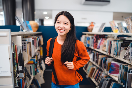 The portrait of a happy young woman holding bag standing in library.