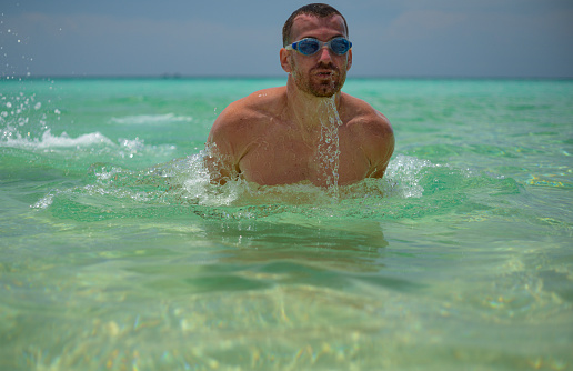 Swimmer athletic man swimming butterfly approaching the camera rising up out of the water. Male athlete fitness model training outside.