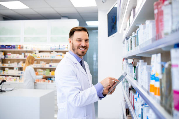 farmacéutico de pie junto a los estantes con medicamentos y escribiendo en tableta en la farmacia. - pill bottle pharmacy medicine shelf fotografías e imágenes de stock