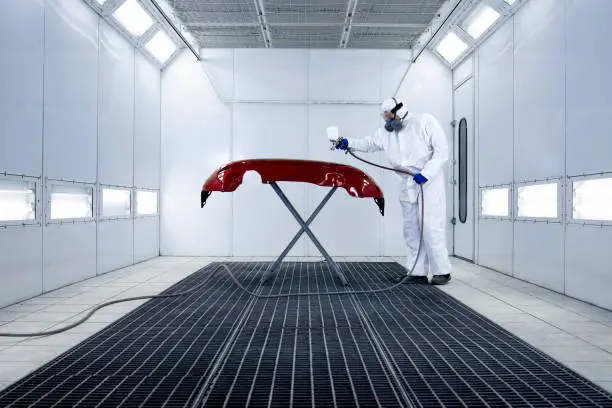 Photo of Car painter in protective clothes and mask painting automobile bumper with metallic paint and varnish in chamber workshop.