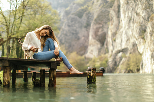 Beautiful woman sitting on the dock and playing with her cat. They are far away from the crowded city, enjoying quiet time with a beautiful calming view