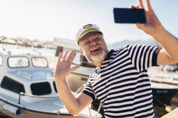 Mature man standing near the sea dressed in a sailor's shirt and hat using smart phone