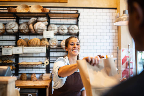 Bakery owner giving food package to customer Bakery owner giving take out food package to customer. Smiling woman working in cafe assisting a customer. bakery photos stock pictures, royalty-free photos & images