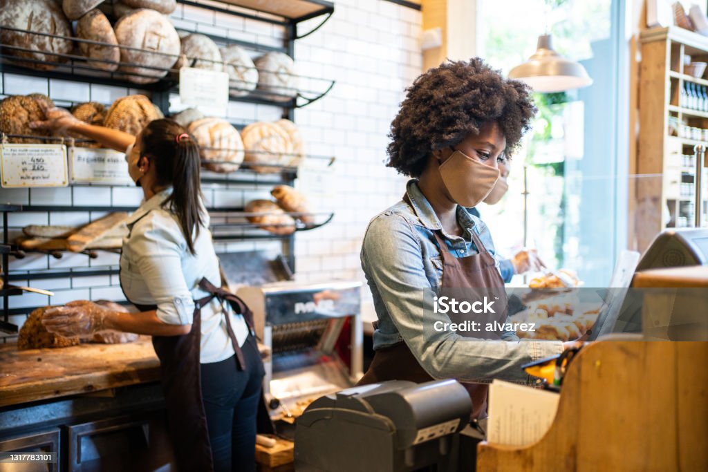 People working in cafe during pandemic wearing face masks African woman working on cash register with colleagues displaying food in rack at background. People working in cafe during pandemic wearing face masks. Bakery Stock Photo