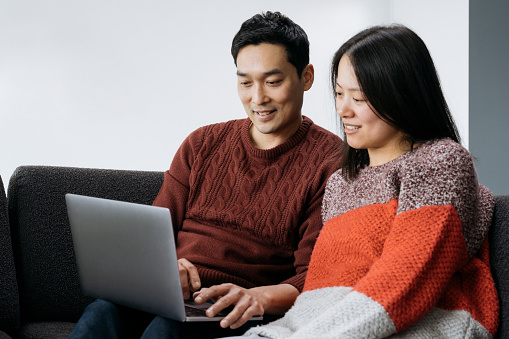 Waist-up view of smiling man and woman in casual clothing sitting next to one another on sofa enjoying weekend entertainment on wireless device.
