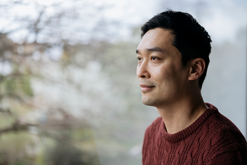 Three-quarter front view of early 40s man with short black hair wearing rust colored pullover sweater and looking away from camera with serene expression.