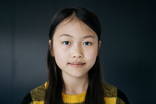Close-up of 13 year old girl with long black hair wearing yellow and black pullover sweater and looking at camera with contented expression.