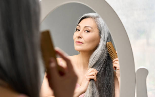matura donna asiatica anziana di mezza età che guarda specchio pettinare i suoi capelli grigi. - beautiful senior woman foto e immagini stock