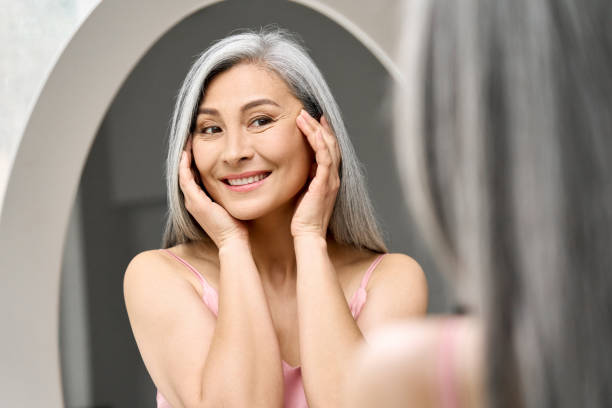 Happy mid aged Asian woman looking at mirror. Antiaging beauty care concept. Happy middle 50 years aged asian woman with gray hair looking at mirror reflection examining touching face enjoying antiaging beauty treatments. Beauty hydrate skin care wrinkle prevention concept. midsection stock pictures, royalty-free photos & images