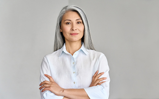 Stylish confident adult 50 years old Asian female psychologist standing arms crossed looking at camera at gray background. Portrait of sophisticated grey hair woman advertising products and services.