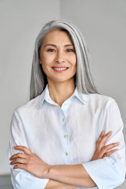 Vertical portrait of mature 50 years Asian business woman on grey background. Smiling confident adult 50 years old Asian female professional standing arms crossed looking at camera at gray background. Portrait of sophisticated grey hair woman advertising products and services. 40 44 years photos stock pictures, royalty-free photos & images
