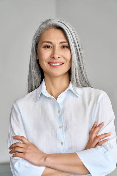 Vertical portrait of mature 50 years Asian business woman on grey background. Smiling confident adult 50 years old Asian female professional standing arms crossed looking at camera at gray background. Portrait of sophisticated grey hair woman advertising products and services. asian woman stock pictures, royalty-free photos & images