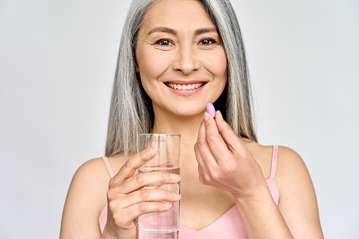 Happy middle aged 50s Asian woman holding pill and glass of water taking dietary supplements. Portrait of smiling adult attractive woman taking care of health in menopause, isolated on white.