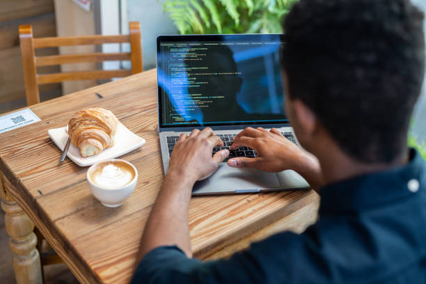 Over the shoulder view of a man working on laptop at cafe. Cup of coffee and croissant on table with man using laptop.