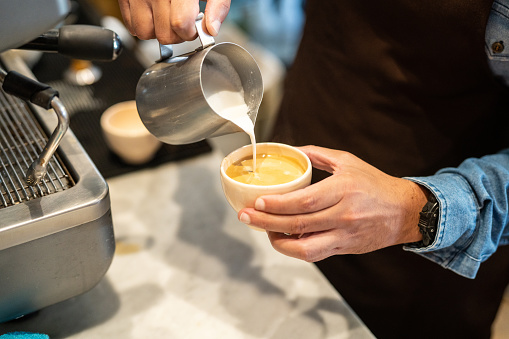 Hand of a barista pouring milk in a coffee latte cup. Closeup of a man making latte art with frothed milk on coffee shop kitchen.