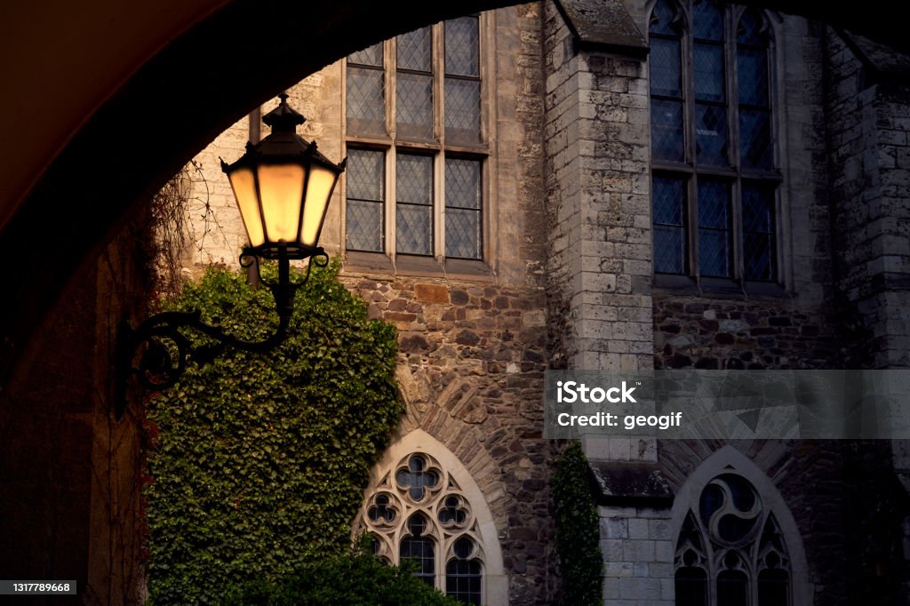 Shining yellow lantern in front of medieval sacral building with high windows and stone walls, view through dark vault Gothic Style Stock Photo