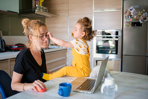 Young mother attending an online meeting in her home kitchen and her adorable baby wanting to play with her mother's glasses. Academician mother with glasses and cute baby girl wearing yellow overalls