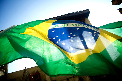 Brazilian flag flying with the blue sky in the background and the sun shining.