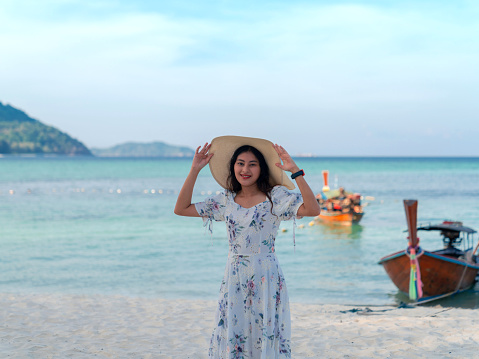 woman in dress holding wide brim hat on head standing at sea view sand beach, blue sea and sky on Lipe islands, Thailand, happy travel, vacation in summer