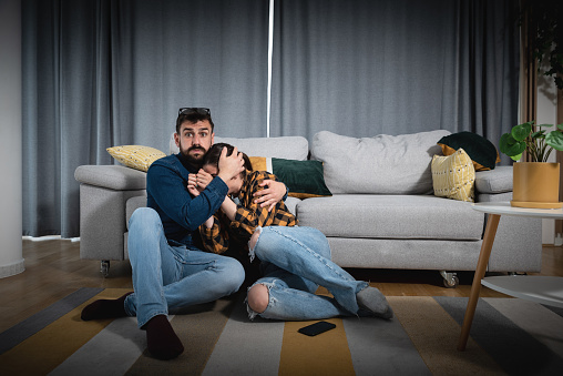 Young couple sitting on the floor of their apartment watching scary horror movie while woman is very scared and man hugs her with her head hidden on his chest