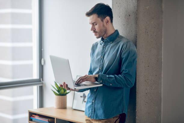 istock Dark-haired man standing near the window and working on a laptop 1317803505