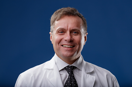 Portrait of a happy smiling middle-aged man in a white lab coat facing the camera over a blue studio background in a concept of medical doctor, technician, or scientist