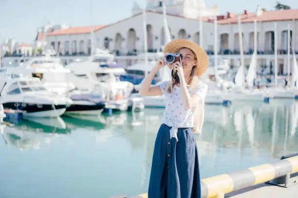A young woman traveler in a straw hat takes a photo on the camera in the seaport.