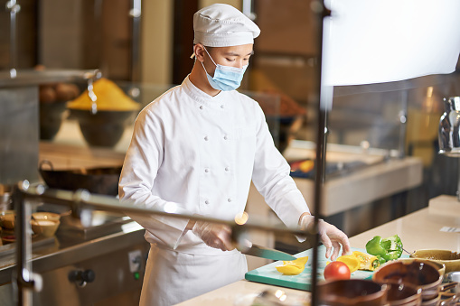 Focused Asian male chef in full uniform and face mask cutting vegetables on a kitchen board