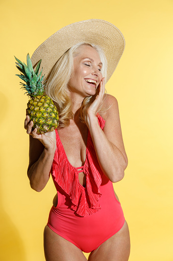 Careless mature blonde woman in red swimsuit and straw hat laughing with eyes closed, holding fresh pineapple fruit while posing isolated on yellow background in studio. People, summer vacation, rest