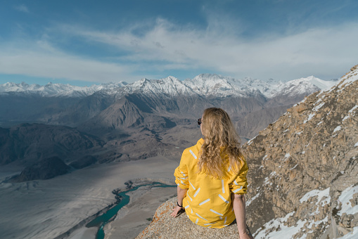 Young Caucasian woman in yellow jacket  sitting on the edge of hanging rock above the river in Northern Pakistan