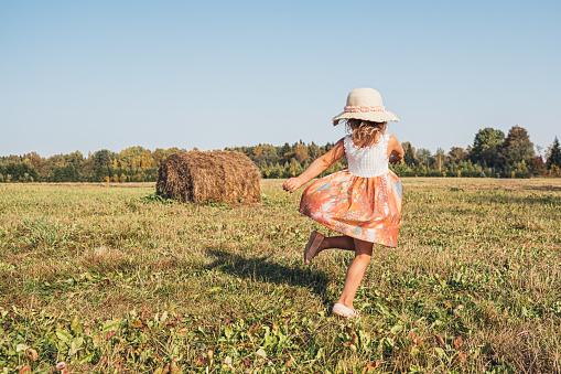 Cute little girl in a straw hat and summer dress fun dancing on the field with a big stack of hay. Summer in the village.