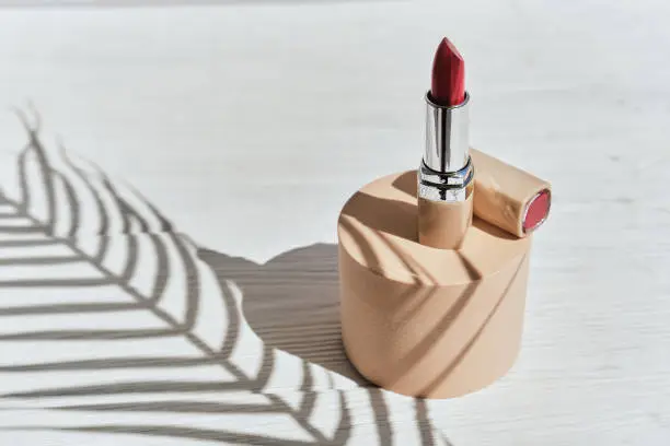 Red lipstick on a beige cylinder podium with shadows from branches of palm on a white background. Trend style. Mockup for the presentation of cosmetics.