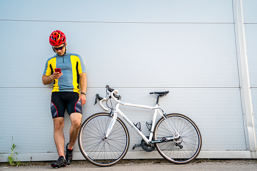 Handsome young cyclist man using mobile phone while standing near his bicycle against the white wall