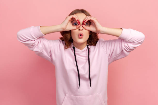 Portrait of amazed teen girl with curly hair in hoodie looking through binoculars gesture and expressing surprise, zooming vision, exploring distance Portrait of amazed teen girl with curly hair in hoodie looking through binoculars gesture and expressing surprise, zooming vision, exploring distance. Indoor studio shot isolated on pink background Distant stock pictures, royalty-free photos & images