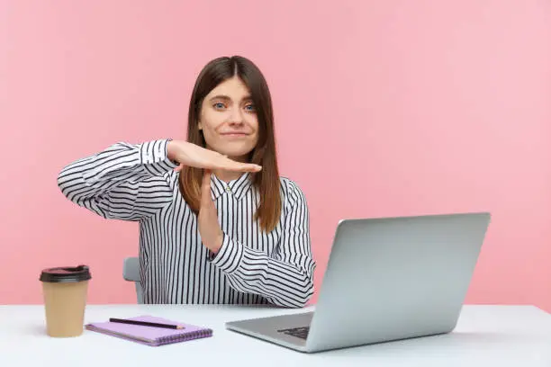 Exhausted overworked woman showing time out gesture, asking for break sitting at workplace with laptop, professional burnout. Indoor studio shot isolated on pink background