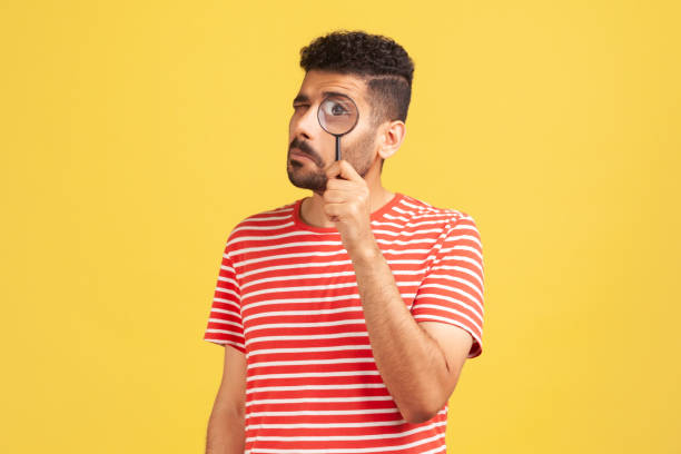 serious bearded man in red striped t-shirt standing, holding magnifying glass and looking at camera with big zoom eye, verifying authenticity - loupe quality control magnification glass imagens e fotografias de stock