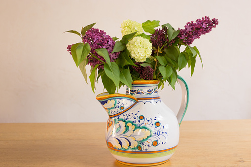 Bouquet of lilacs and snowball viburnum in an Italian majolica pitcher on a wood table.