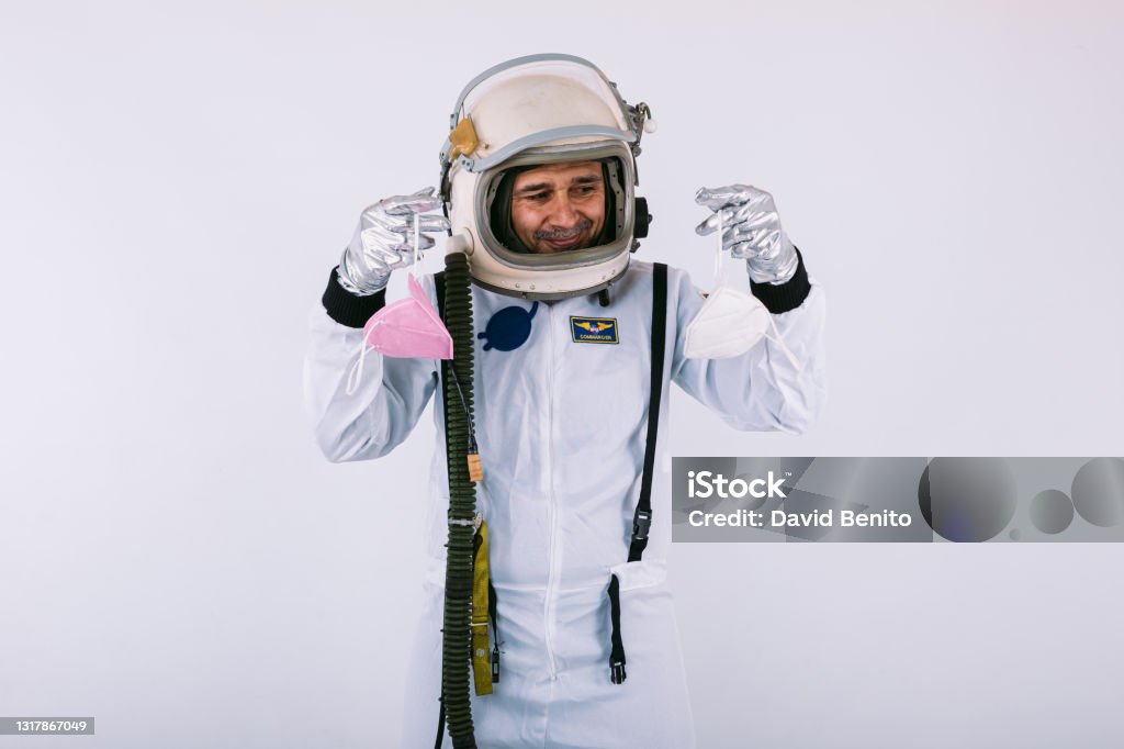 Male cosmonaut in spacesuit and helmet, holding two FPP2 masks, on white background. Covid-19 and virus concept Indoors Stock Photo