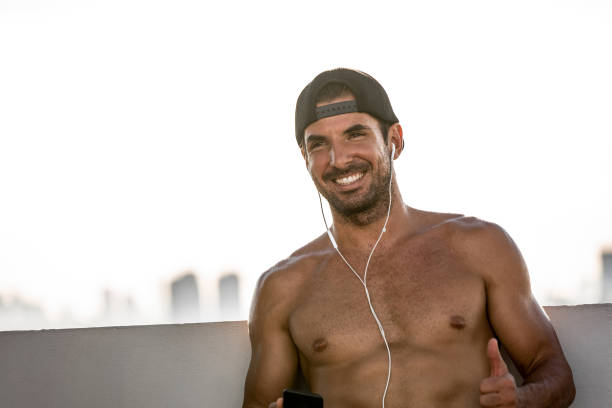 Shirtless smiling handsome sports man wearing earphones listening to music from mobile phone relaxing after workout in open air Shirtless smiling handsome sports man wearing earphones listening to music from mobile phone relaxing after workout in open air happ stock pictures, royalty-free photos & images