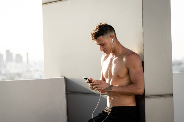 Shirtless handsome sports man wearing earphones listening to music from mobile phone relaxing after workout on rooftop in evening sunlight Shirtless handsome sports man wearing earphones listening to music from mobile phone relaxing after workout on rooftop in evening sunlight happ stock pictures, royalty-free photos & images