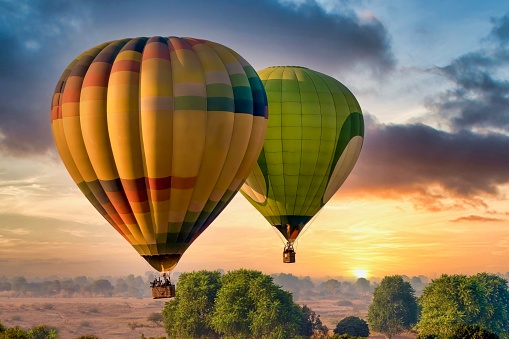 Two colorful hot air balloons, their baskets filled with unidentifiable tourists, drift above the rural landscape of fields and trees, while the sun rises on the horizon.