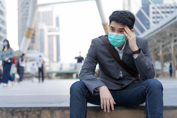 nahaufnahme junge hispanische mitarbeiter mann tragen maske und sitzen mit kopfschmerzen gefühl vor dem gebäude mit überfüllten menschen für gesundheitsversorgung und umweltverschmutzung konzept - quit scene stock-fotos und bilder