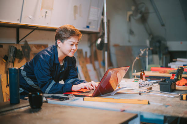 soudeur mûr asiatique de femme mûr travaillant dans le sourire d’atelier utilisant la dactylographie d’ordinateur portatif - se courber en avant photos et images de collection