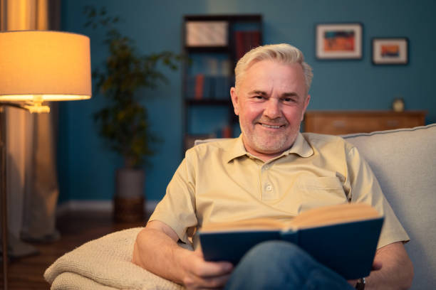istock Portrait of a smiling, contented elderly man sitting on a couch looking into a camera. Elderly man holding a book, an old book. Grey haired guy with stubble contentedly sitting in living room on couch 1317914672