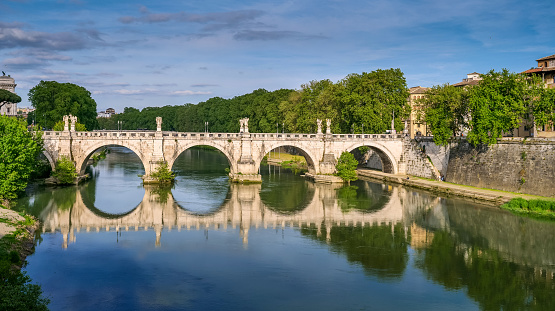 A suggestive warm light envelops the bank of the Tiber river and the baroque Ponte Sant'Angelo, in the historic center of Rome, near the Vatican area, creating a pictorial effect with reflections on the water. Image in high definition format.