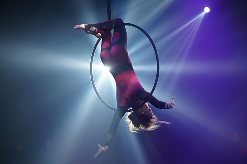Flexible woman gymnast make acrobatic element upside down on aerial hoop backlit in white purple lights , circus show concept