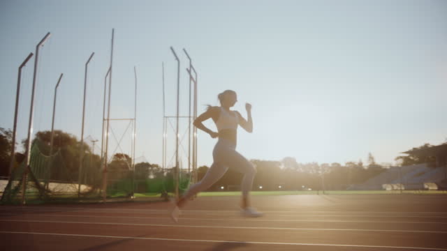 Beautiful Female Athlete in Light Blue Sports Top and Leggings Running Extremely Fast in an Outdoors Stadium. She is Sprinting on a Warm Summer Afternoon. Finess Woman Doing Her Jogging Training.