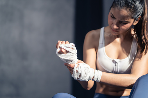 Portrait of Asian active beautiful athlete trainer girl wrapping exercise glove for workout exercise at fitness gym. Sportswoman in sportswear suffer from pain in hand and massaging wrist in stadium.