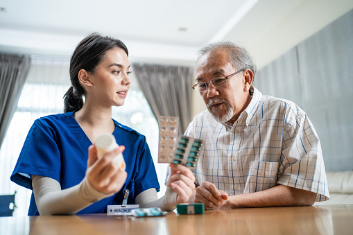 El paciente masculino mayor asiático consulta con la enfermera médica en la atención del hogar de ancianos. Cuidador terapeuta farmacéutico chica sostener píldoras de medicamentos botella, explicar la prescripción a un hombre discapacitado mayor en c photo
