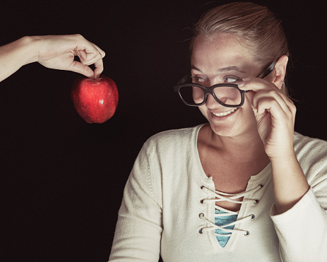 Mid adult woman looks pleased to be given an apple by the disembodied hand of a young person.